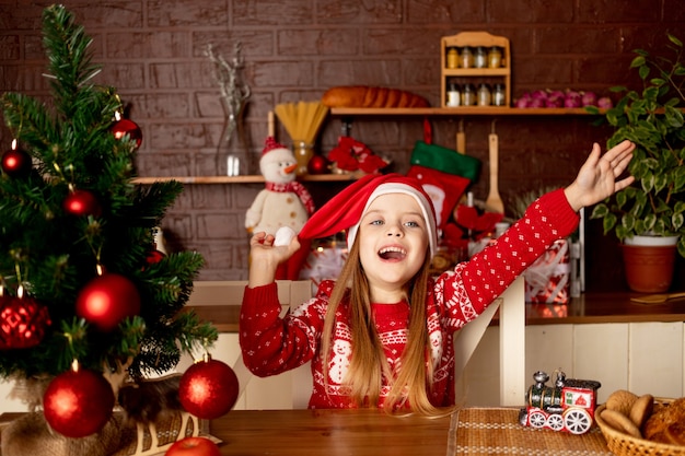 Una niña niño feliz con un gorro de Papá Noel en una cocina oscura en el árbol de Navidad con bolas rojas se regocija y sonríe, el concepto de año nuevo y Navidad