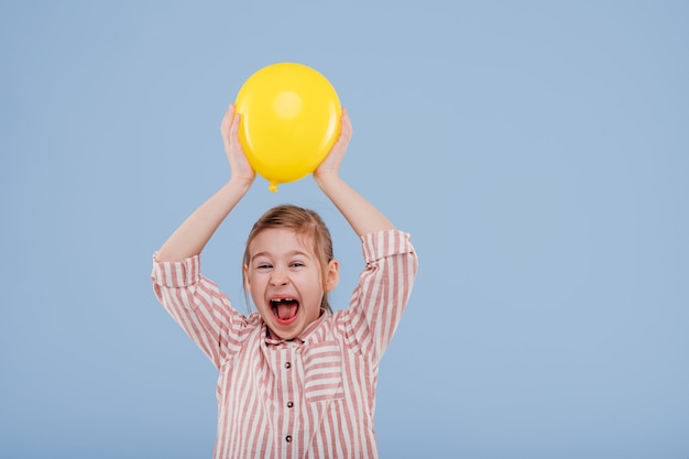 Niña niño feliz sostiene globo amarillo vestido con camisa a rayas aislado sobre fondo azul copia s ...