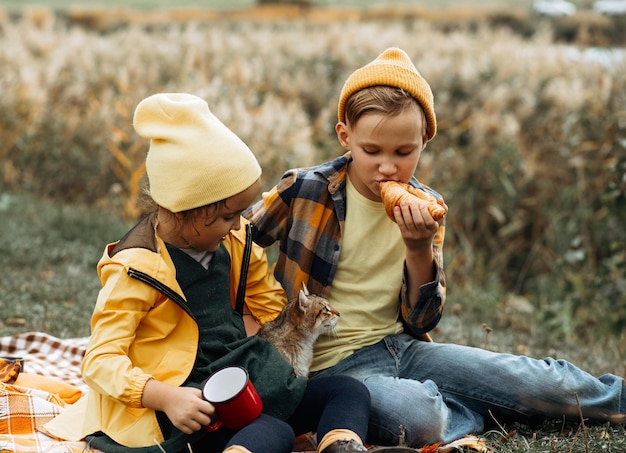 Niña y niño lindos y sonrientes sentados cerca de un estanque o lago en cuadros comiendo croissant y bebiendo cacao Niño descansando con su familia en la naturaleza Picnic Temporada de otoño