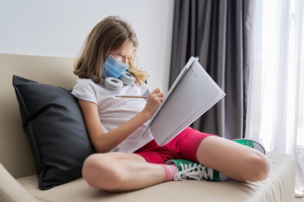 Niña niño en una mascarilla protectora médica sentado en el sofá en casa, un estudiante que estudia con un cuaderno escolar