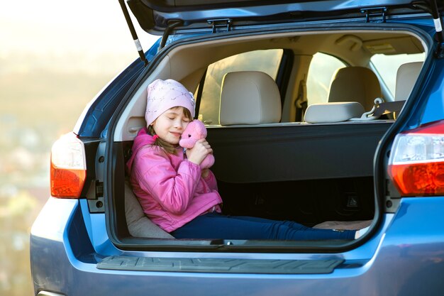 Niña niño muy feliz jugando con un oso de peluche de juguete rosa en el maletero de un coche.