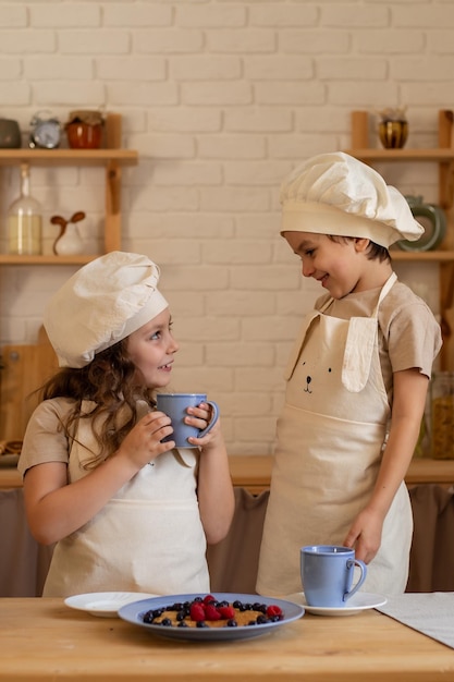 una niña y un niño con sombreros y delantales de chef beben bebidas con panqueques y bayas se miran el uno al otro