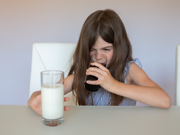 Una niña no quiere beber leche, pero elige un refresco de coca cola. Concepto de alimentación saludable y no saludable