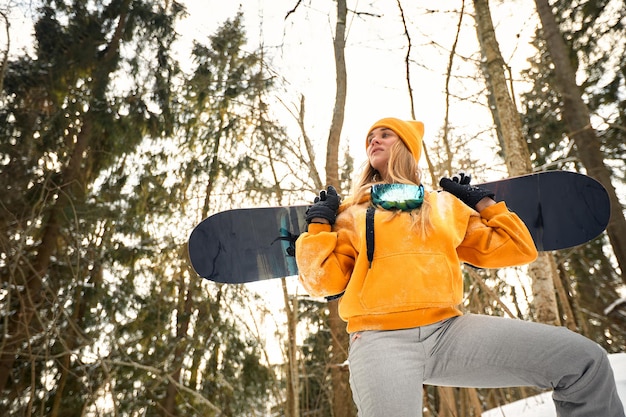 Niña o mujer snowboarder entra para deportes de invierno en el bosque nevado que se encuentra en la nieve y sostiene una tabla de snowboard