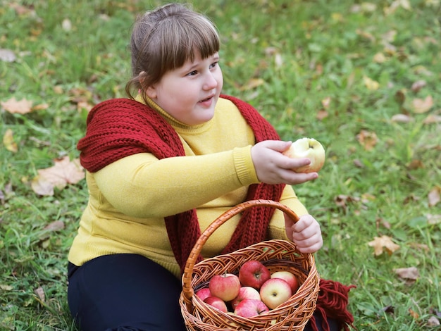 Niña obesa con pañuelo rojo se sienta en la hierba y sostiene la canasta con manzanas en otoño