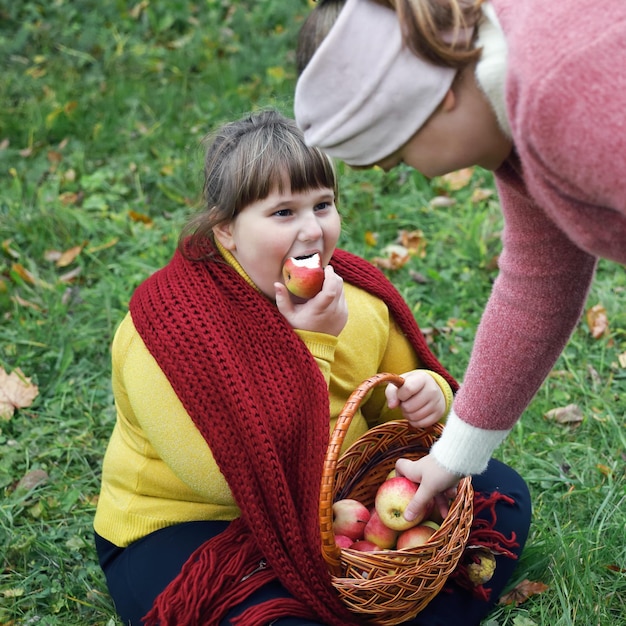 Niña obesa con pañuelo rojo se sienta en la hierba y sostiene la canasta con manzanas en otoño