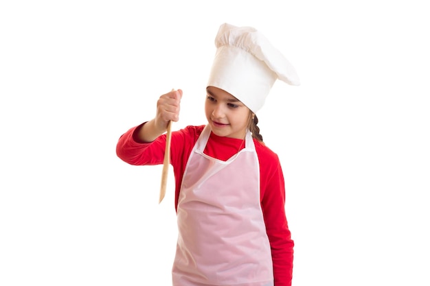 Niña optimista con camisa roja con delantal blanco y sombrero con espátula de cocina en el estudio