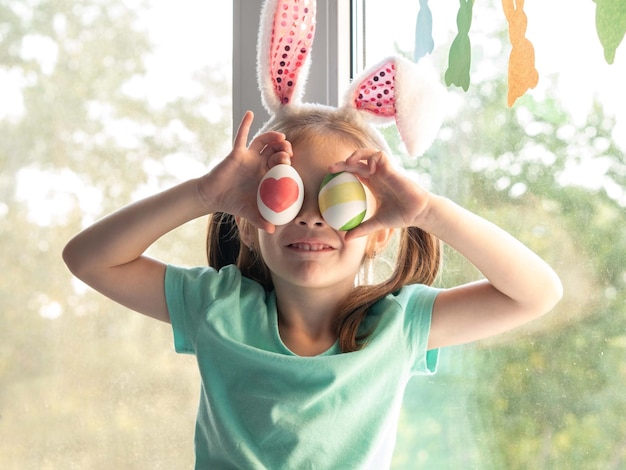 Una niña con orejas de conejo en el alféizar de la ventana pone huevos de Pascua en sus ojos