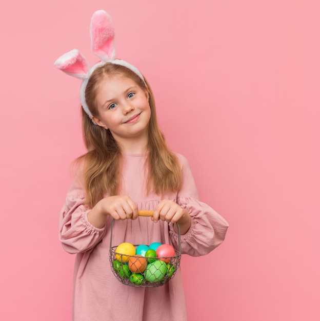 Niña con orejas de conejo y canasta con huevos de pascua sobre fondo rosa