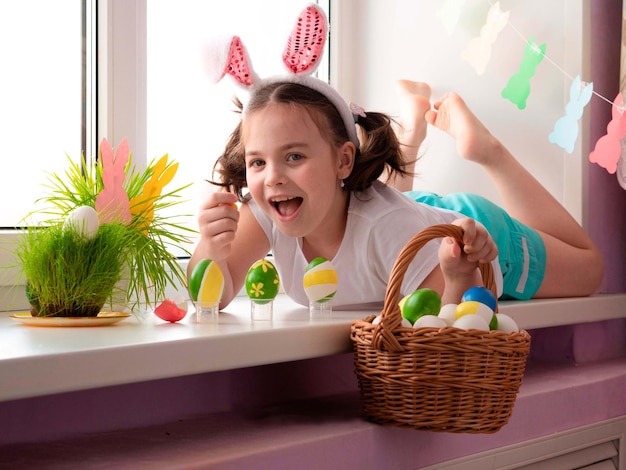 Niña con orejas de conejo yace en el alféizar de la ventana feliz sostiene una canasta con huevos de Pascua