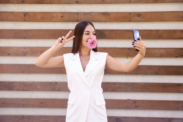 Niña oriental sonriente vestida de blanco con una flor rosa en la boca tomando selfies