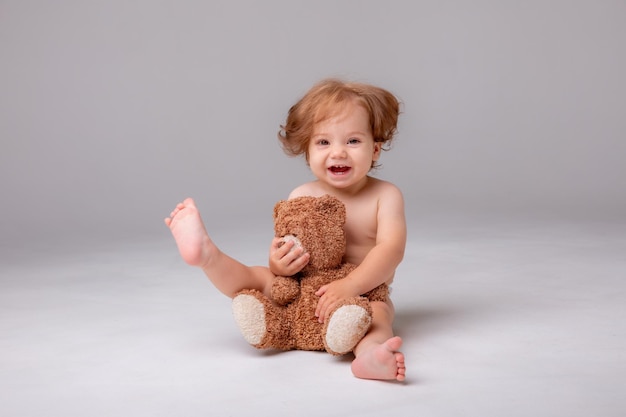 Una niña en un pañal se sienta y juega abrazos con un oso de peluche sobre un fondo blanco.