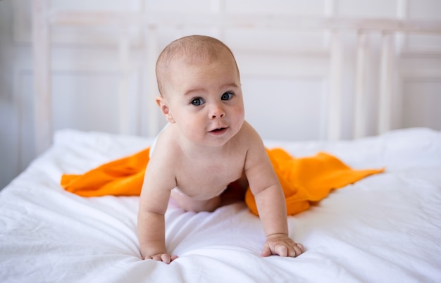 Niña en pañal y con una toalla naranja se arrastra sobre la cama. Desarrollo infantil