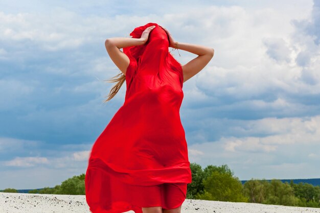 Una niña con un paño rojo en la arena posa para un fotógrafo con el telón de fondo de un cielo nublado. Tela roja en el viento abraza la figura de la niña.