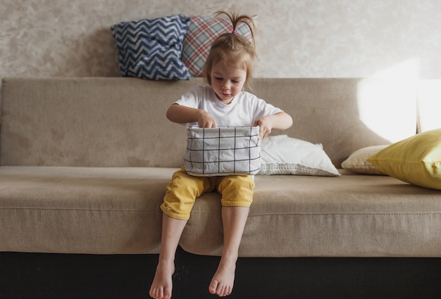 Una niña con pantalones amarillos y una camiseta blanca se sienta en el sofá y dobla cuidadosamente su ropa en una caja de almacenamiento. Asistente de mamá.