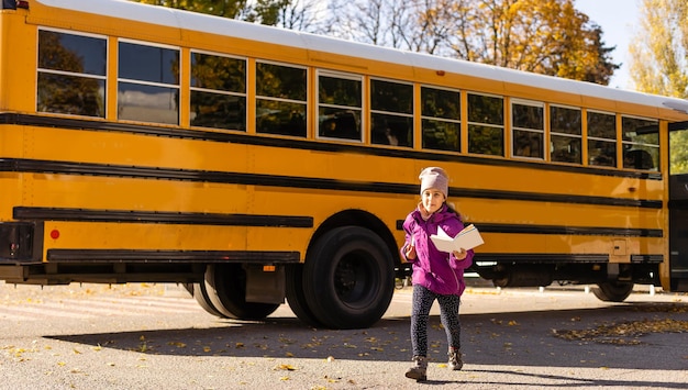 Niña parada junto a un gran autobús escolar con sus libros.