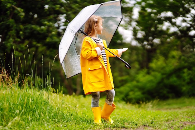 Niña con paraguas transparente jugando bajo la lluvia en el sol Hermosa niña en un manto amarillo.