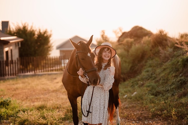 Una niña con parálisis cerebral con un vestido ligero y un sombrero blanco se ríe de un caballo marrón.