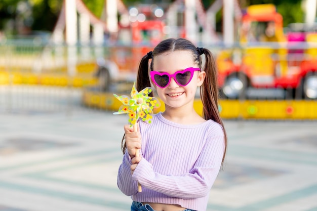 Una niña en un parque de atracciones en verano con un juguete giratorio en las manos sonríe con felicidad cerca de los carruseles con gafas de sol el concepto de vacaciones de verano y vacaciones escolares