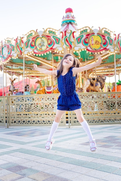 Una niña en un parque de atracciones en verano salta de felicidad alrededor de los carruseles y se ríe del concepto de vacaciones de verano y vacaciones escolares