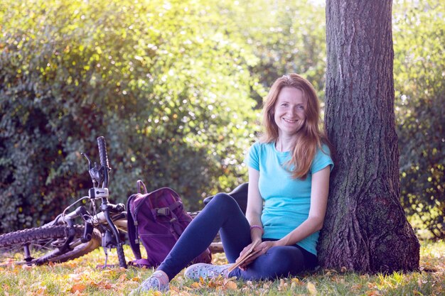 Niña en el parque se sienta debajo de un árbol y lee un libro. al lado hay una bicicleta y una mochila