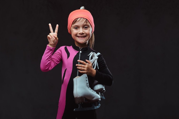 Una niña patinadora feliz vestida con ropa deportiva sostiene patines de hielo en un hombro y muestra la mano de la victoria. Aislado en un fondo de textura oscura.