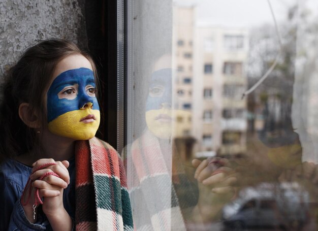 Una niña patriota ucraniana con la bandera de Ucrania en la cara reza sentada en la ventana Los niños están en contra de la guerra en Ucrania