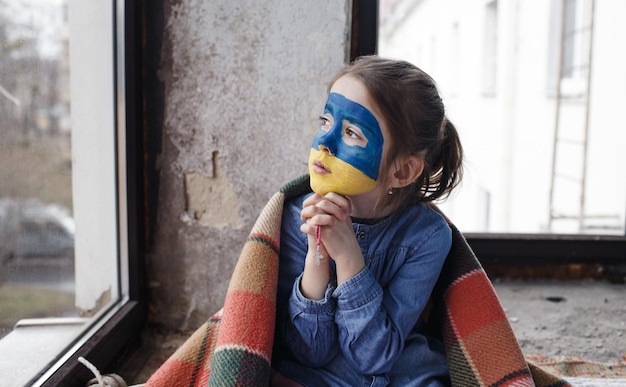 Una niña patriota ucraniana con la bandera de Ucrania en la cara reza sentada en la ventana