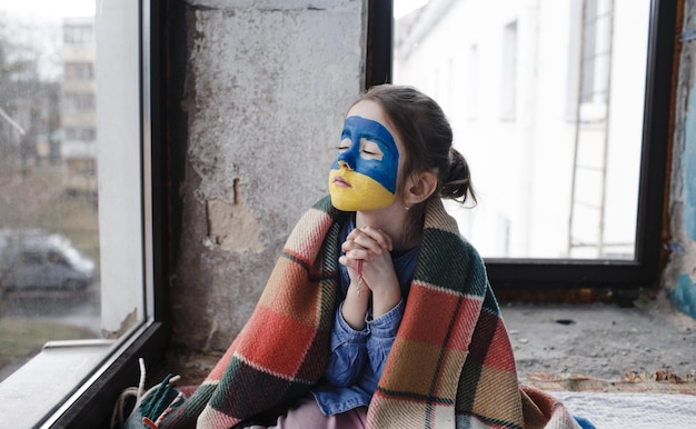 Foto una niña patriota ucraniana con la bandera de ucrania en la cara reza sentada en la ventana