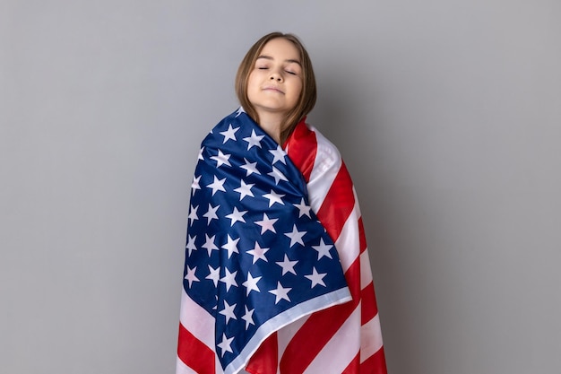 Niña patriótica de pie envuelta en la bandera americana manteniendo los ojos cerrados reubicándose en Estados Unidos