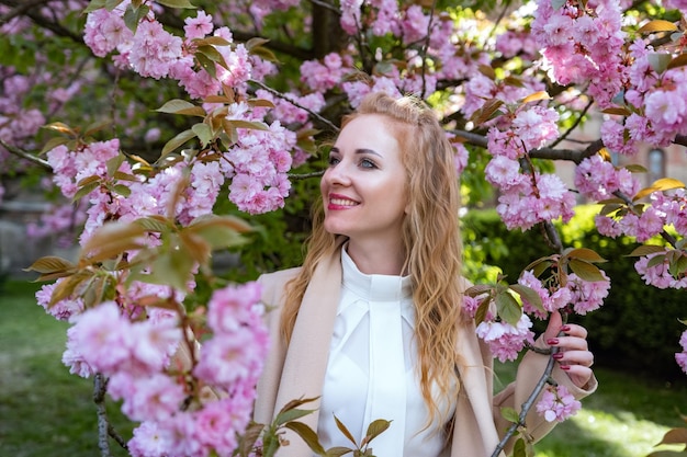 Niña pelirroja con camisa blanca de pie cerca de sakura en un clima soleado de primavera Retrato de una mujer atractiva con cabello ondulado en un parque en flor