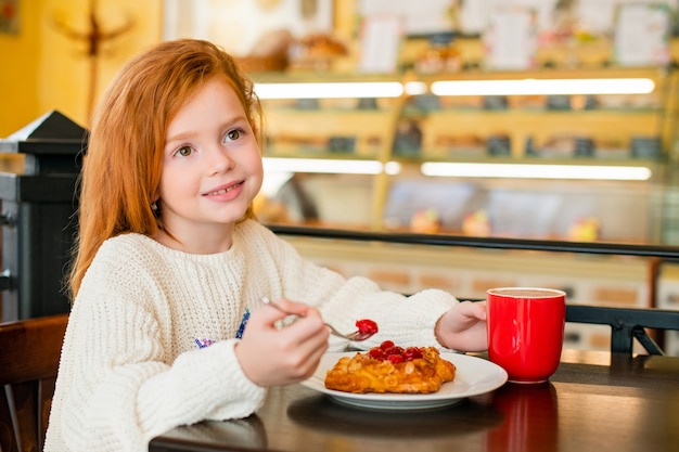 Niña pelirroja comiendo pastel en un café, sonriendo mirando a lo lejos.