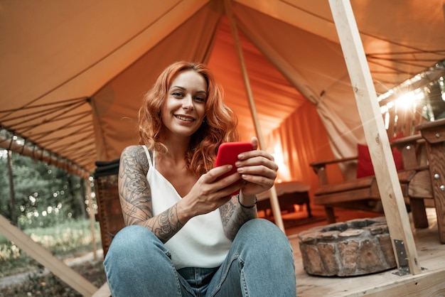 La niña pelirroja sonriente se sienta junto a la carpa y charla con la transmisión de mensajes en el teléfono inteligente, publicando, dando me gusta en las redes sociales. Viaja fuera de la ciudad en el bosque. Cámping.