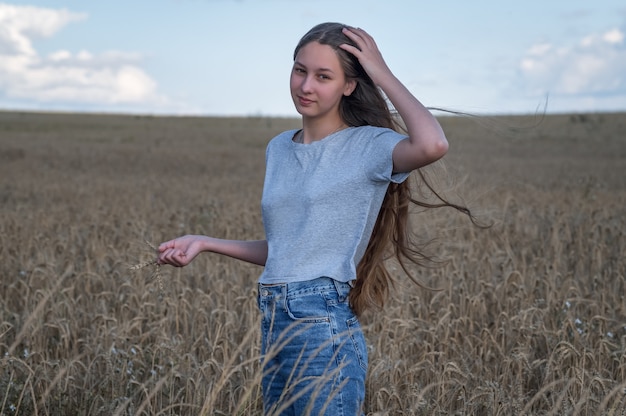 Una niña con el pelo largo en jeans y una camiseta está de pie en un campo de luz natural de centeno