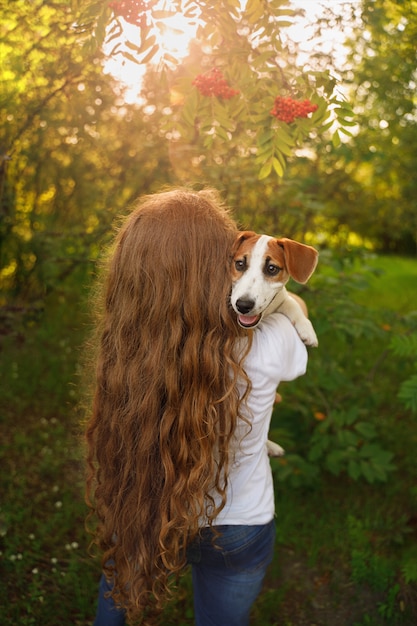 Una niña con el pelo largo y rizado está de pie con la espalda y sosteniendo un cachorro en sus brazos.