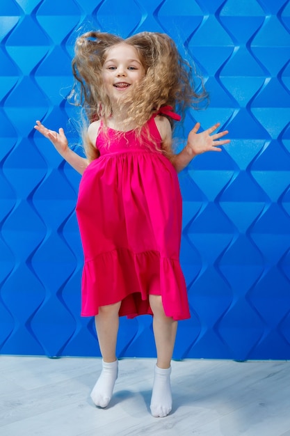 Foto niña con pelo largo y rizado con un vestido rosa sobre un fondo azul baila y se ríe, las emociones de alegría de los niños brillantes, infancia feliz