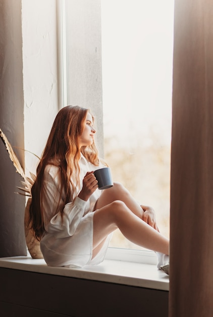 Una niña con el pelo largo se sienta en el alféizar de una ventana con una taza en sus manos