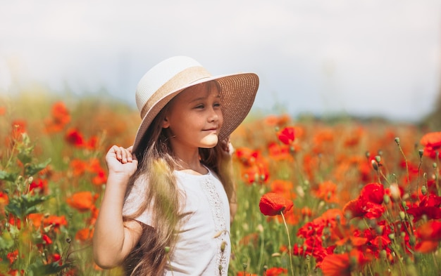 Niña de pelo largo con sombrero posando en el campo de amapolas con el sol de verano