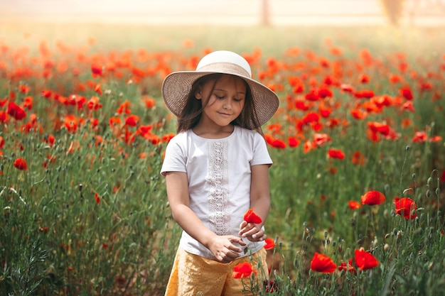 Niña de pelo largo con sombrero posando en el campo de amapolas con el sol de verano