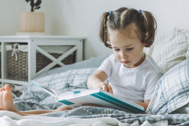 Niña de pelo oscuro 2-4 con dos colas de caballo se sienta en la cama con almohadas y lee el libro