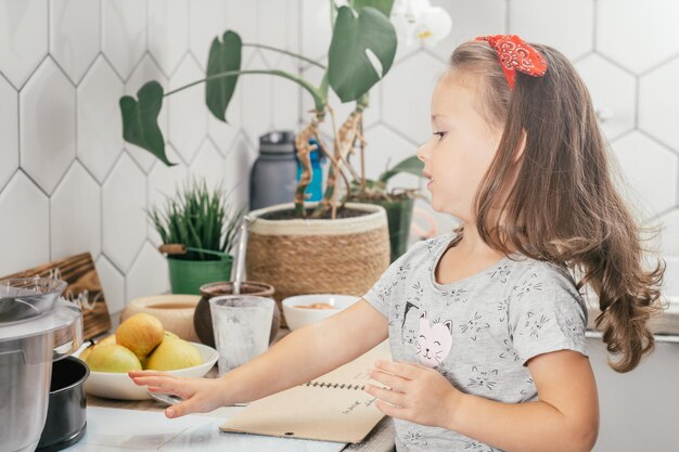 Niña de pelo oscuro de 3 años con diadema hornea pastel de manzana, se encuentra cerca de la mesa con ingredientes, utensilios y libro de recetas.