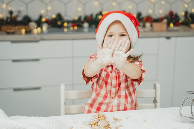 Niña de pelo oscuro de 3 años con gorro de Navidad rojo y camisa a cuadros prepara masa para galletas de jengibre en cocina decorada de Navidad blanca con guirnaldas de luces. Feliz navidad, año nuevo