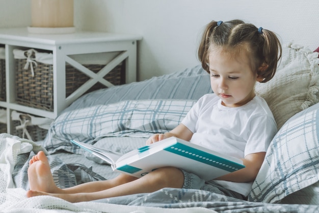 Niña de pelo oscuro con dos colas de caballo se sienta en la cama con almohadas y lee el libro