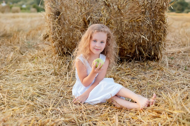 una niña de pelo rizado está sentada junto a un pajar en un campo con una manzana verde en las manos