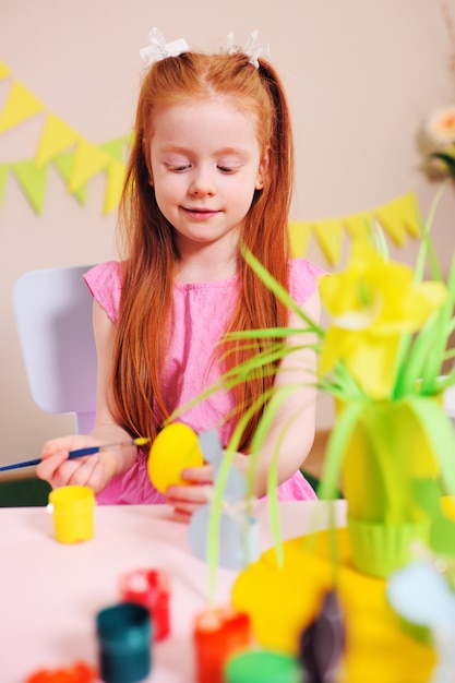 Niña con el pelo rojo para colorear un huevo amarillo sobre la mesa. Decoración de pascua