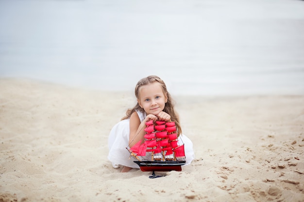 Niña con el pelo rubio en el vestido blanco que sostiene la nave con las velas escarlatas. Esperando amor, soñando concepto. Niño sentado en la orilla del mar, sostiene un velero de juguete en sus manos. Concepto de viaje y aventura