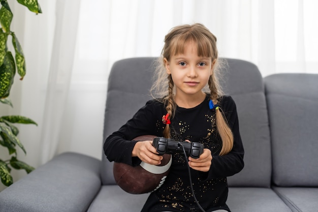 niña con pelota de rugby y joystick para jugar videojuegos en casa