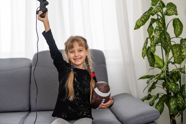 niña con pelota de rugby y joystick para jugar videojuegos en casa