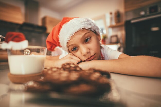 Una niña pensativa mira en una taza de leche y un plato con galletas y esperando a Santa Claus.