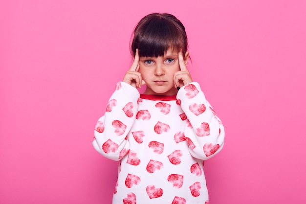 Niña pensativa mirando a cámara, piensa en cosas importantes, manteniendo los dedos cerca de las sienes, siendo pensativa y concentrada, aislada sobre la pared rosa.